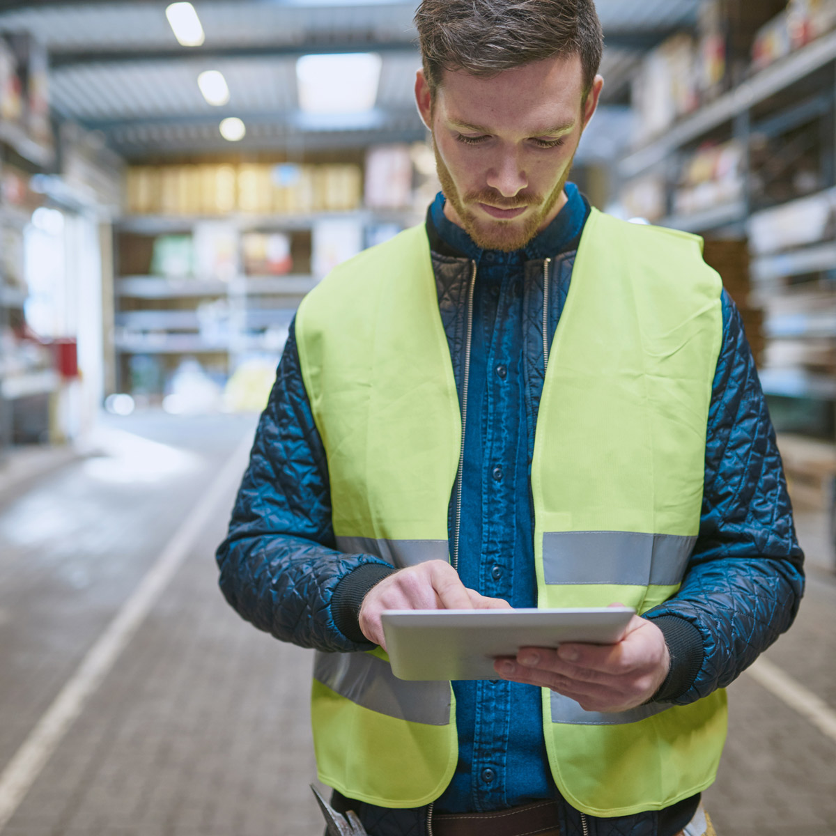 Man in high visibility vest reviewing a tablet inside a warehouse.