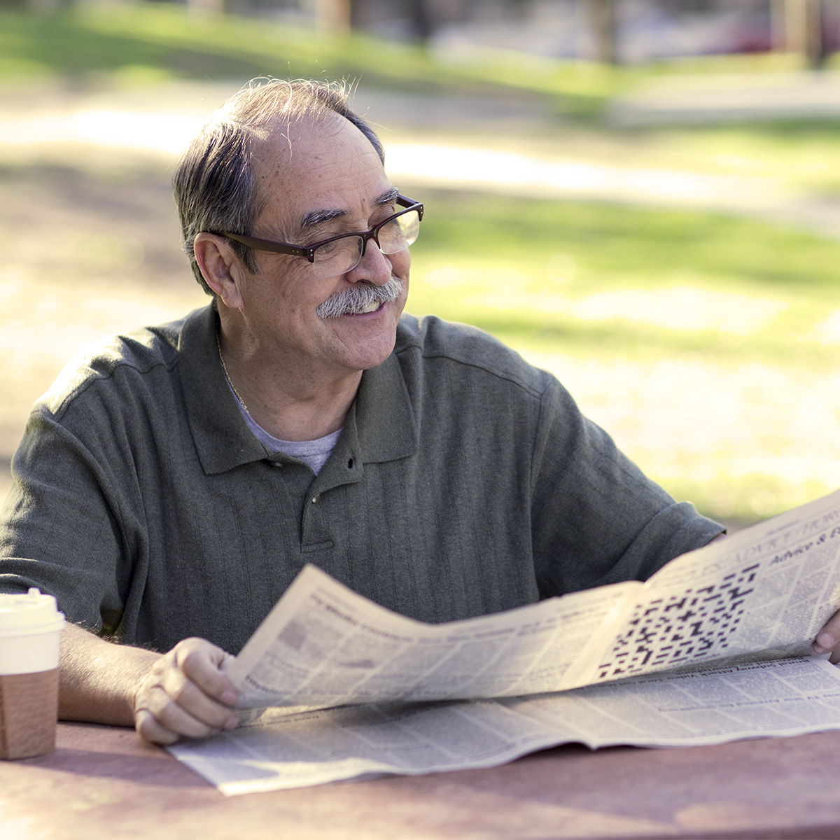 A man enjoying coffee and reading the newspaper in the park on a nice day.