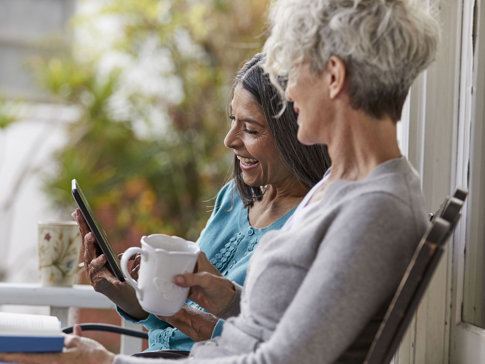 Two laughing women gaze into a tablet as they sit outside and enjoy their morning coffee.