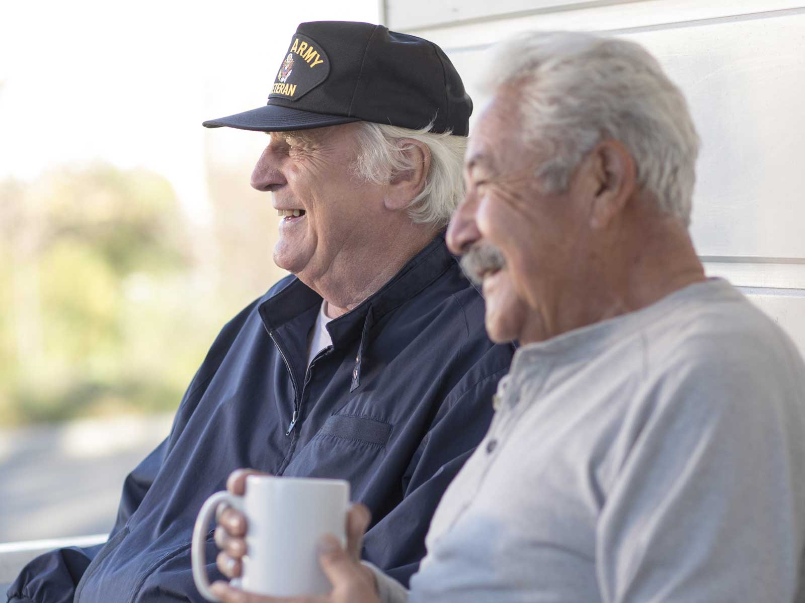 2 older men smiling and drinking coffee, 1 wearing a hat that reads “US Army Veteran.”