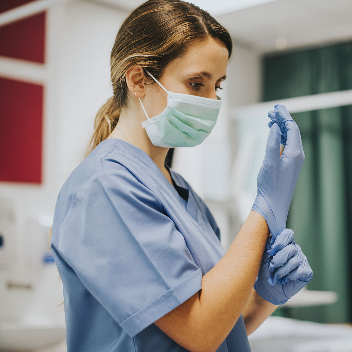 Medical worker in mask and scrubs puts on surgical gloves.
