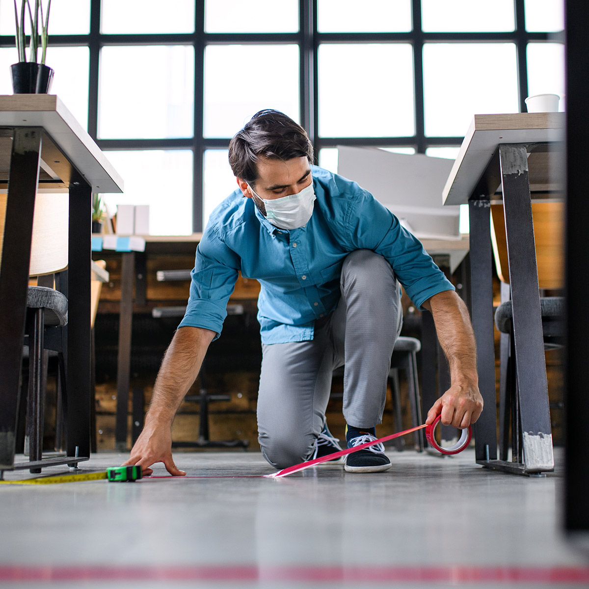 Man wearing a mask crouches on the floor while measuring and marking the distance between tables.