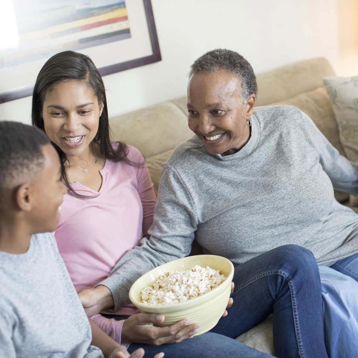 Una abuela, hija y dos nietos riendo y disfrutando un momento juntos, sentados en el sofá con un recipiente con palomitas de maíz.