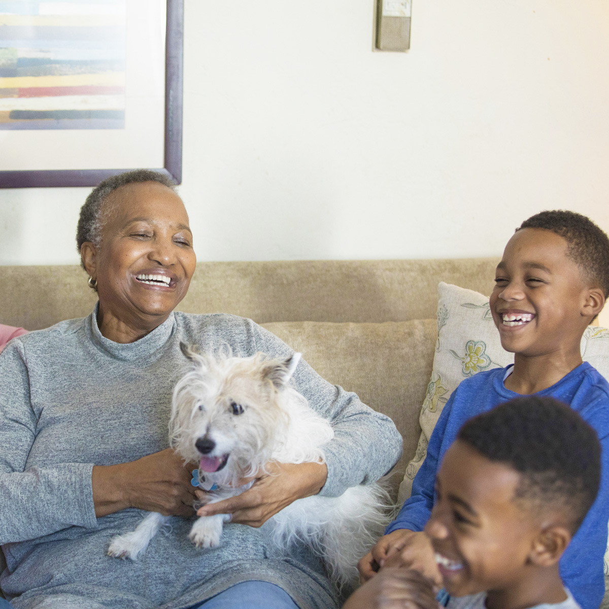 A grandmother on the couch smiling and laughing with her two grandsons while petting a small dog on her lap.