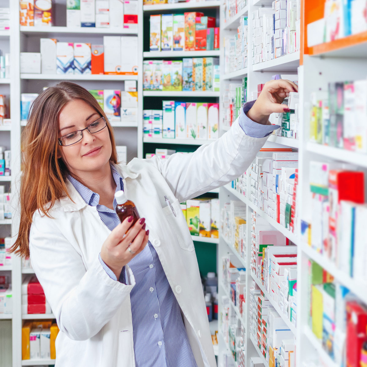 A pharmacist reading the back of a prescription bottle while she reaches for another prescription from the pharmacy shelf.