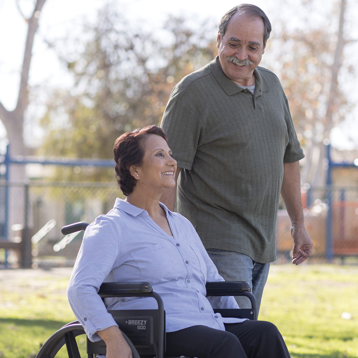 A man and a woman in a wheelchair take a walk through the park