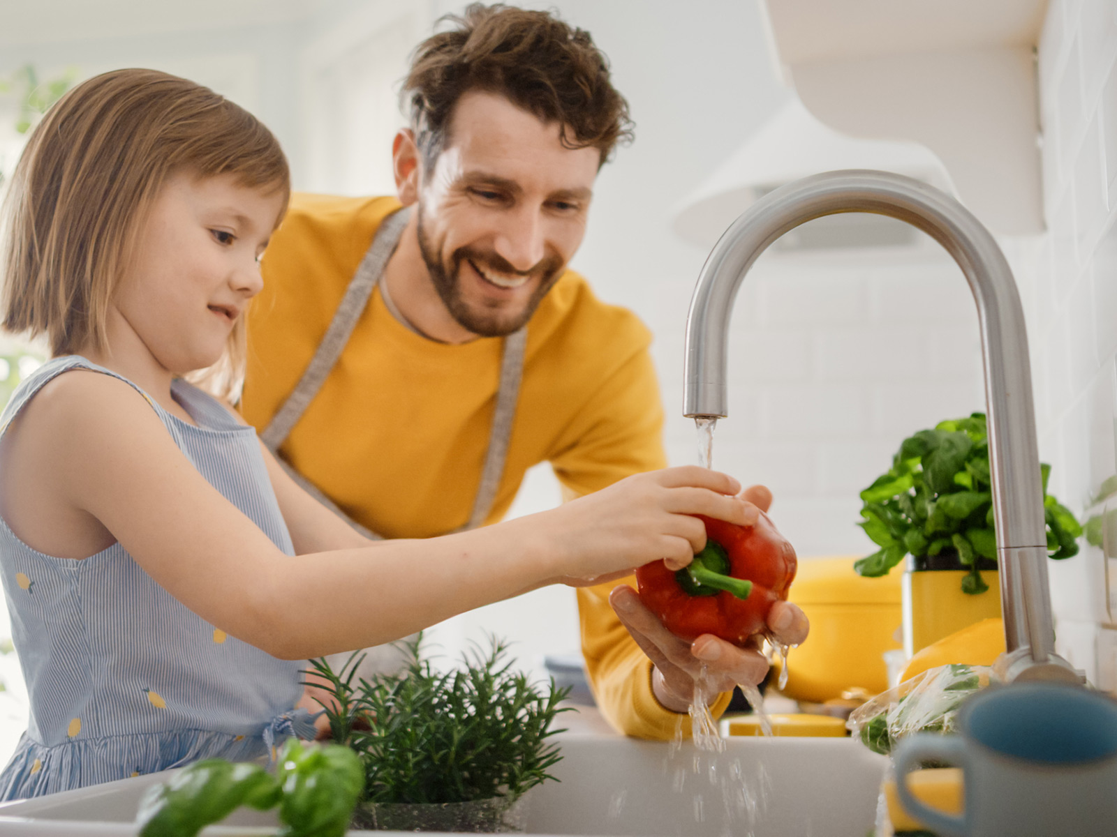 Father smiles while he helps young daughter clean vegetables in the kitchen sink