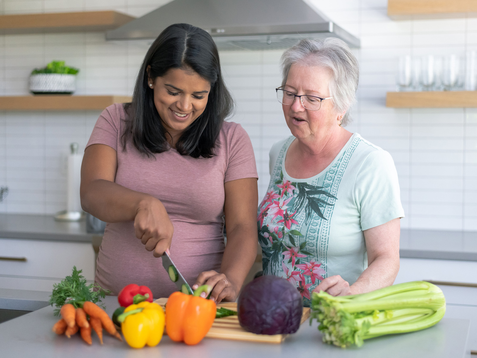 Pregnant woman chops vegetables on kitchen counter as older woman looks on