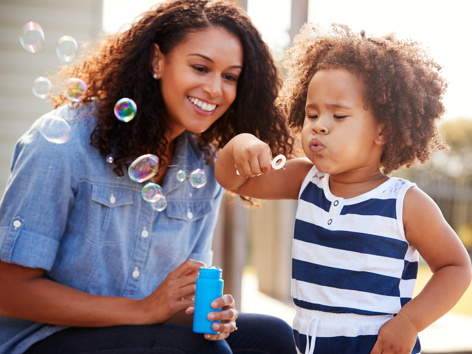 Young child blows bubbles outside while mother looks on