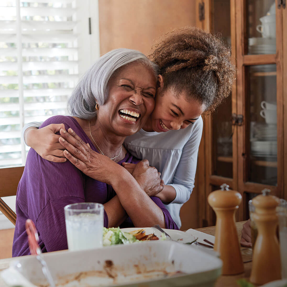 A grandmother and her granddaughter laugh and hug at the dinner table.