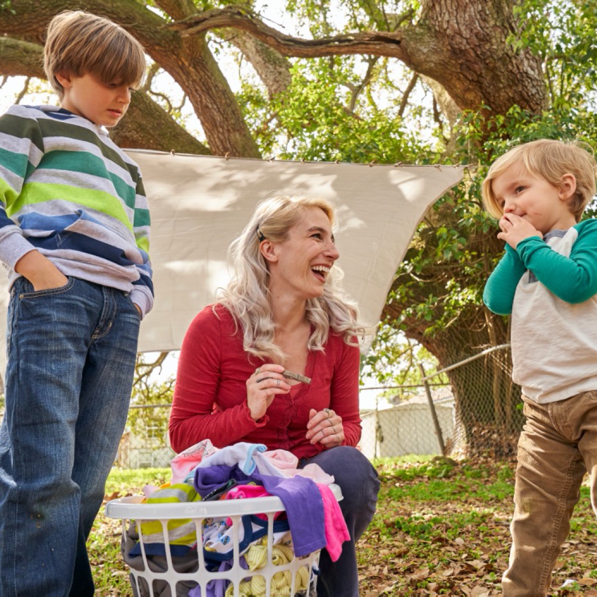Mother and her two sons outside pulling laundry off the line and placing it in the hamper on the ground.