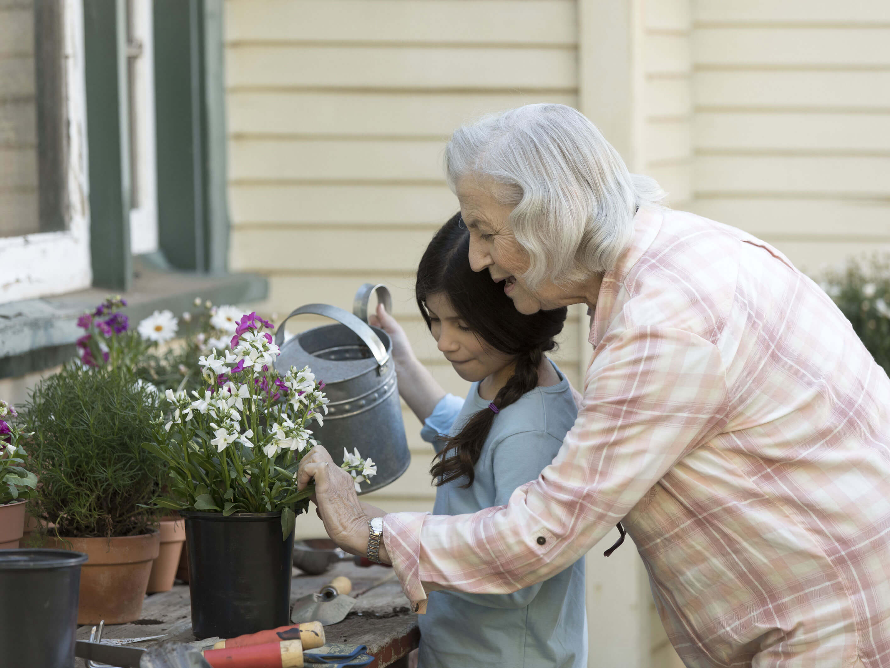 A senior woman helps her young granddaughter water a pot of flowers sitting on a table outside of her home.