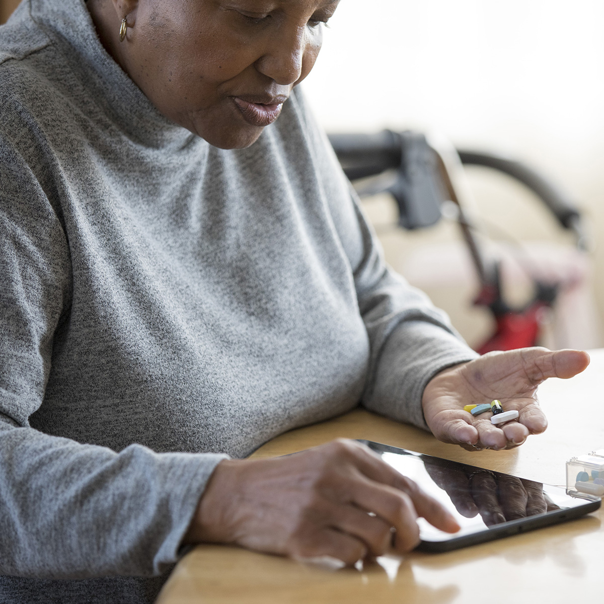 Una mujer busca información en su tableta, sentada a la mesa de la cocina con un medicamento en la mano.