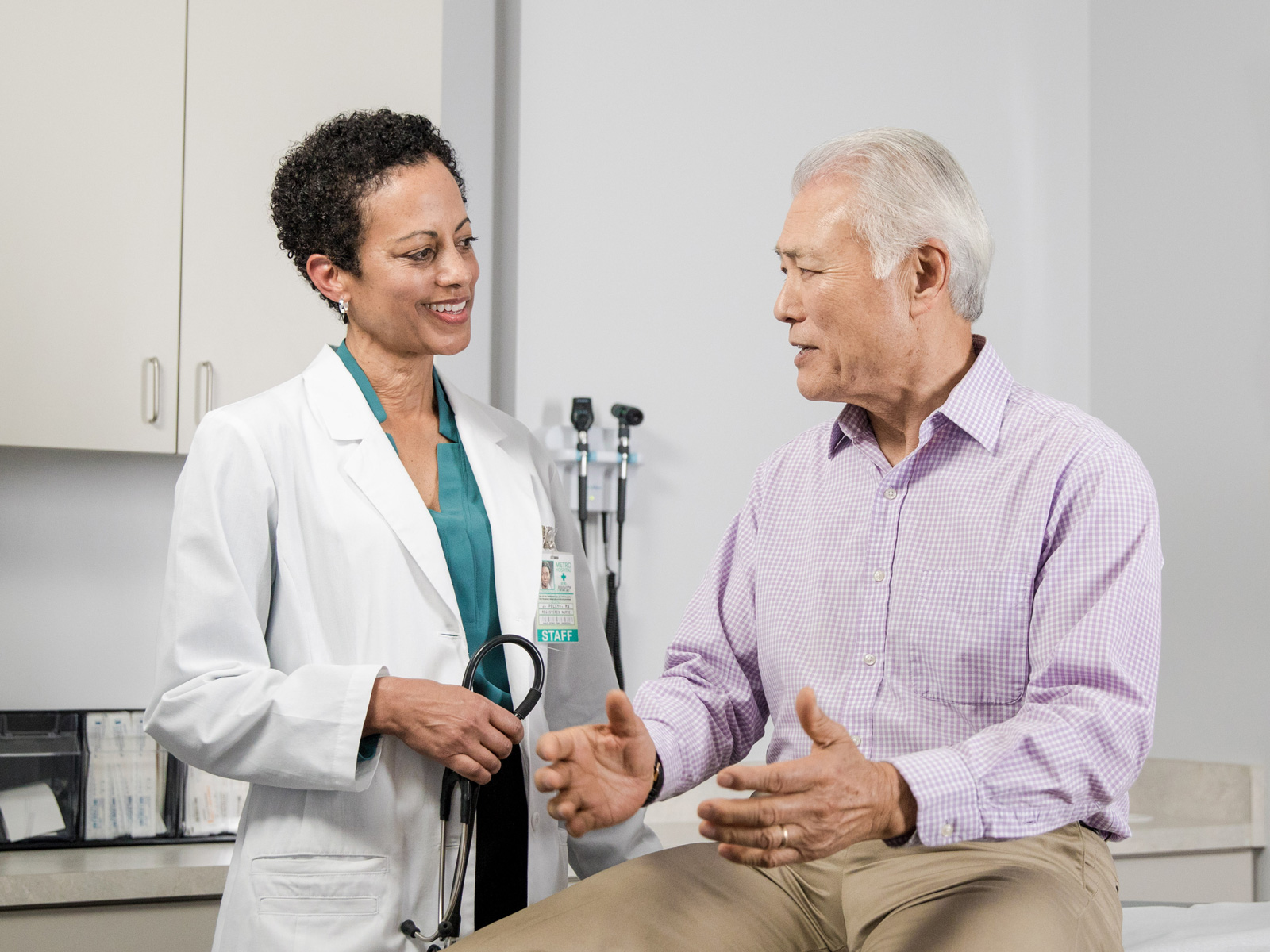 A doctor holding a stethoscope consults her older male patient in an exam room.